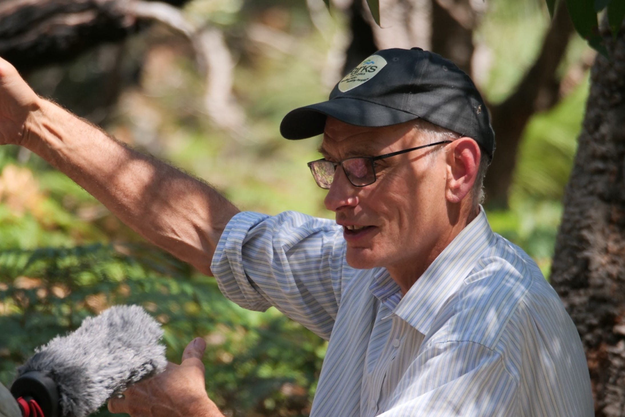 Mark Norman is pointing to something as he stands in the shade of a vegetated area.