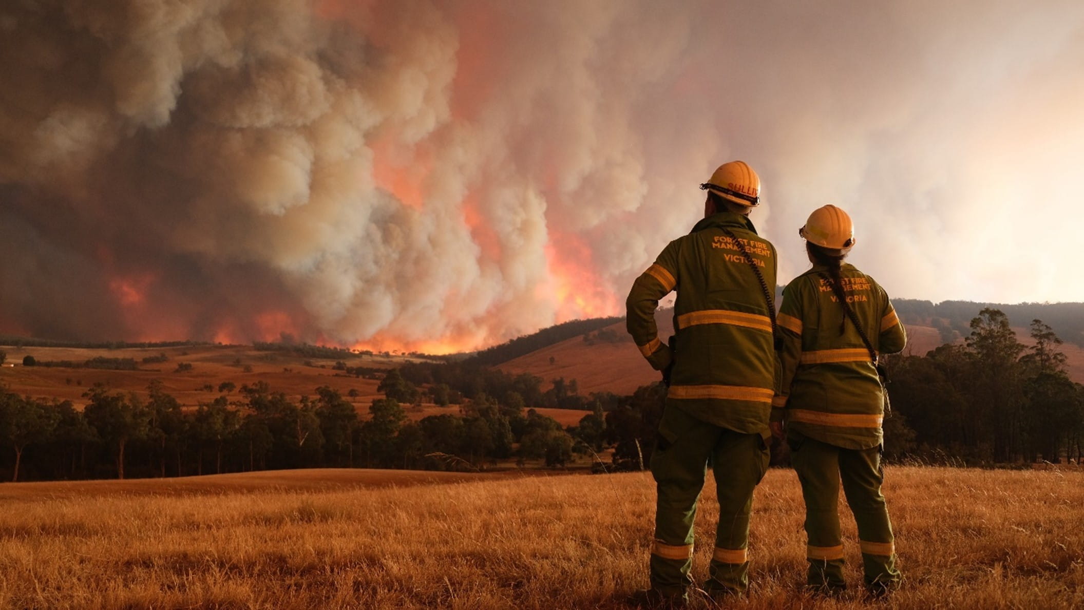 Two firefighters stand looking at a humungous fire enevloping the landscape. Smoke is billowing out of the fire.