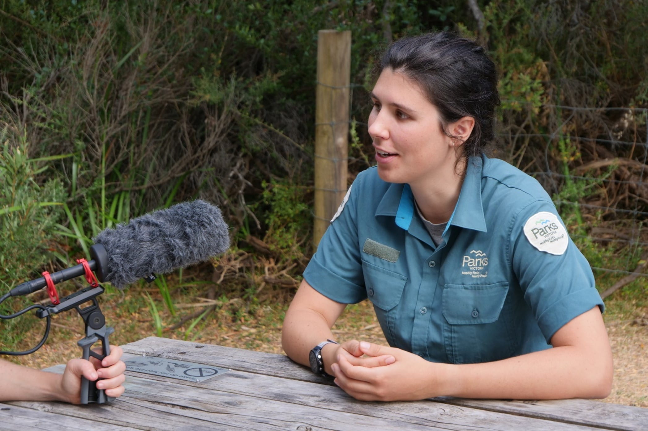 Ruby Wicks sits at a table and speaks into a grey microphone. 