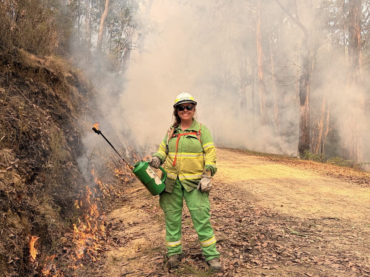 Parks Victoria ranger Hailey Braggins wearing a fire management uniform and doing a controlled burn on a dirt road in the forest. She's smiling and surrounded by smoke.