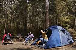 A man talks to his wife who is sitting on a camp chair as he exists his tent.