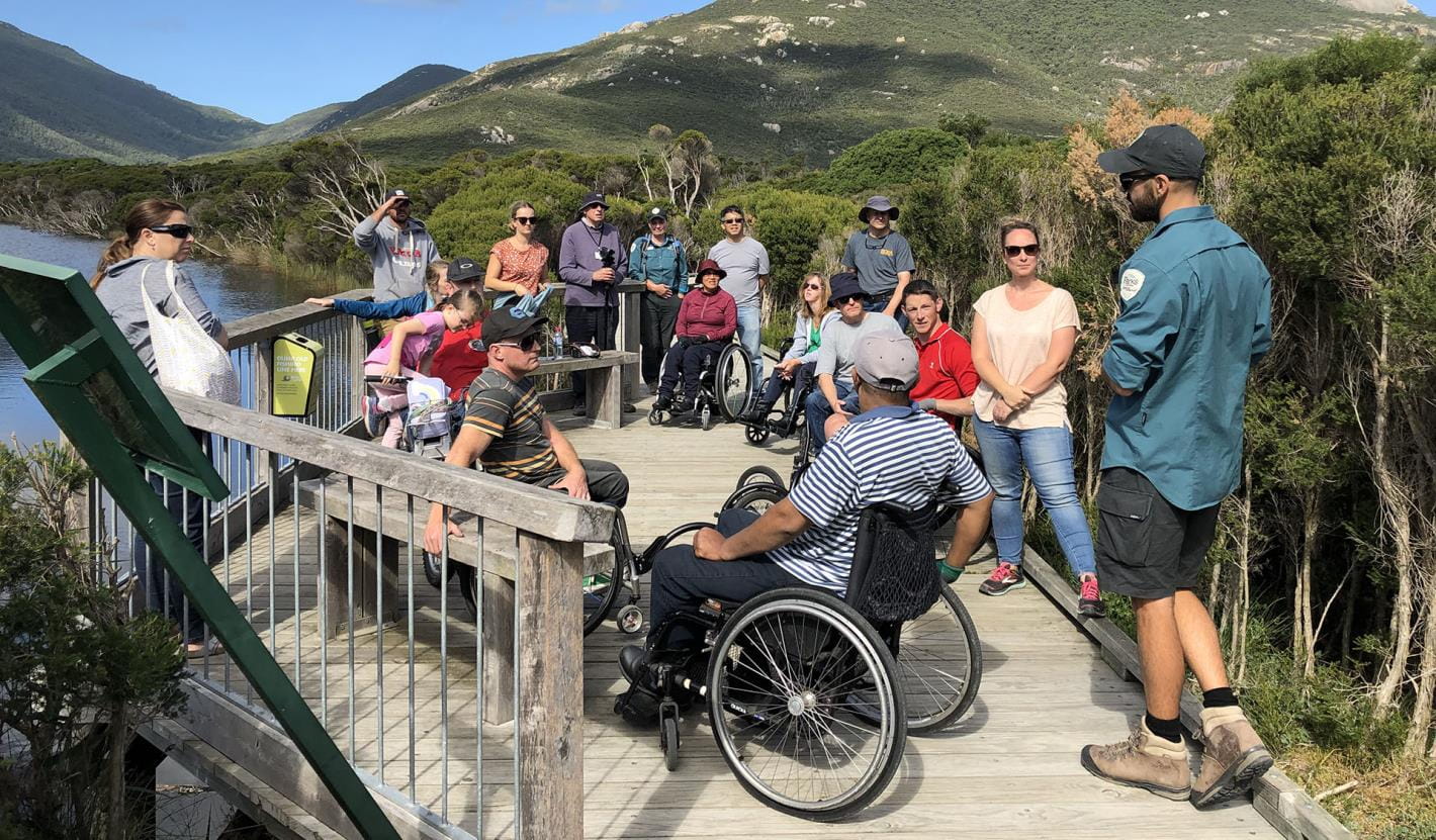 A group of people with various physical abilities gathered with rangers on a boardwalk beside a river with bushland and a mountain in the background.
