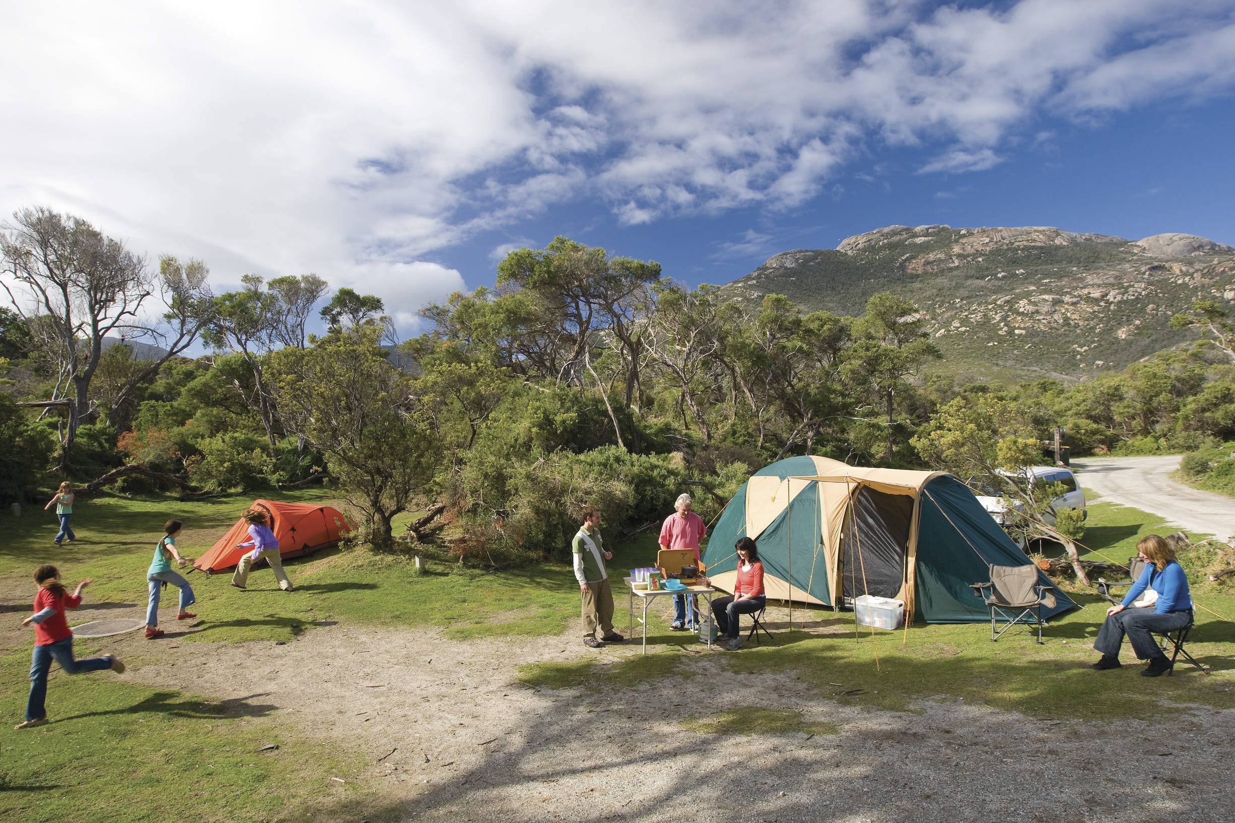 Two tents with people sitting and playing in the foreground. Mountain and blue sky in background