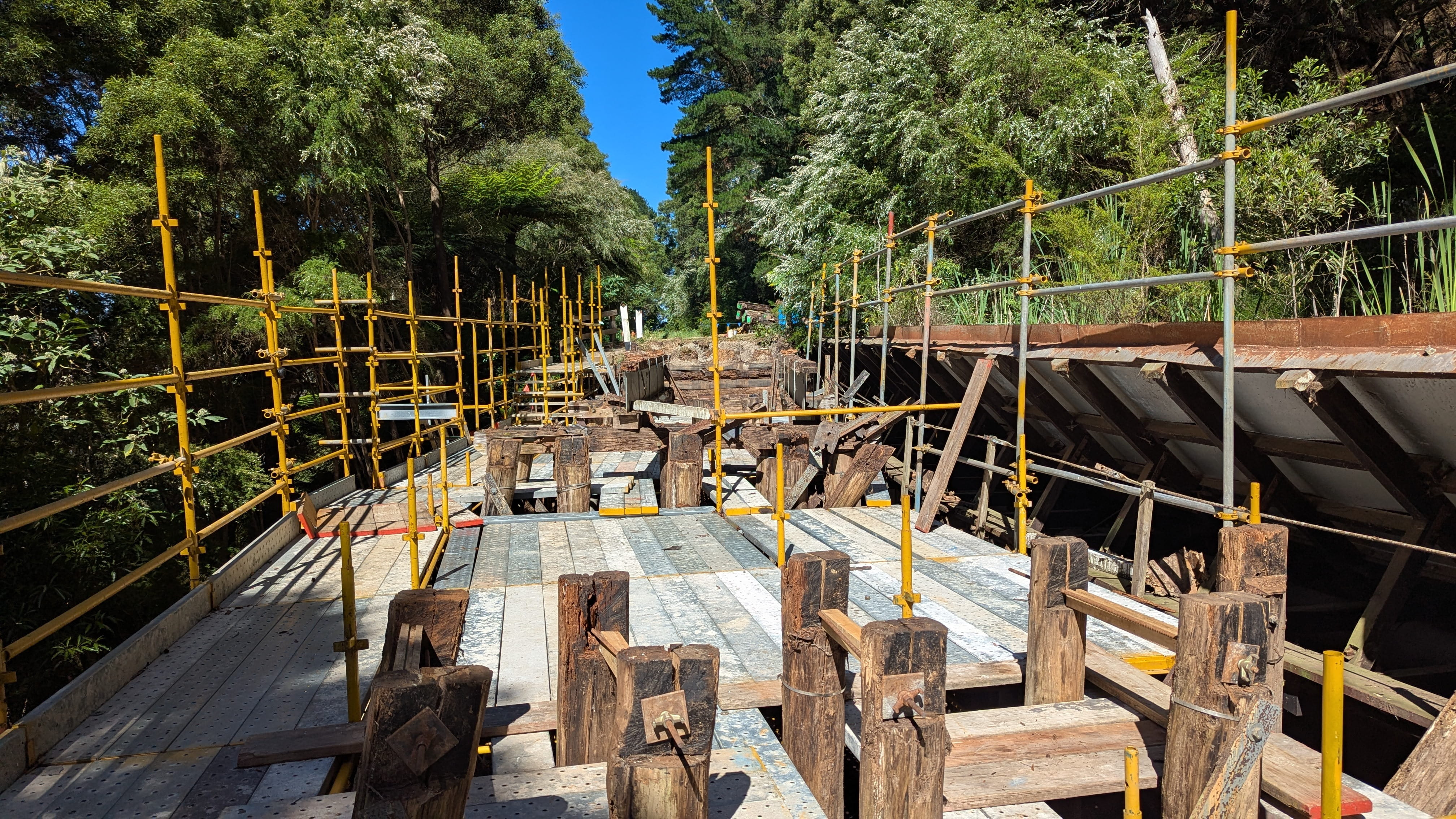 Construction of new bridge with timber posts and scaffolding surrounded by green vegetation.