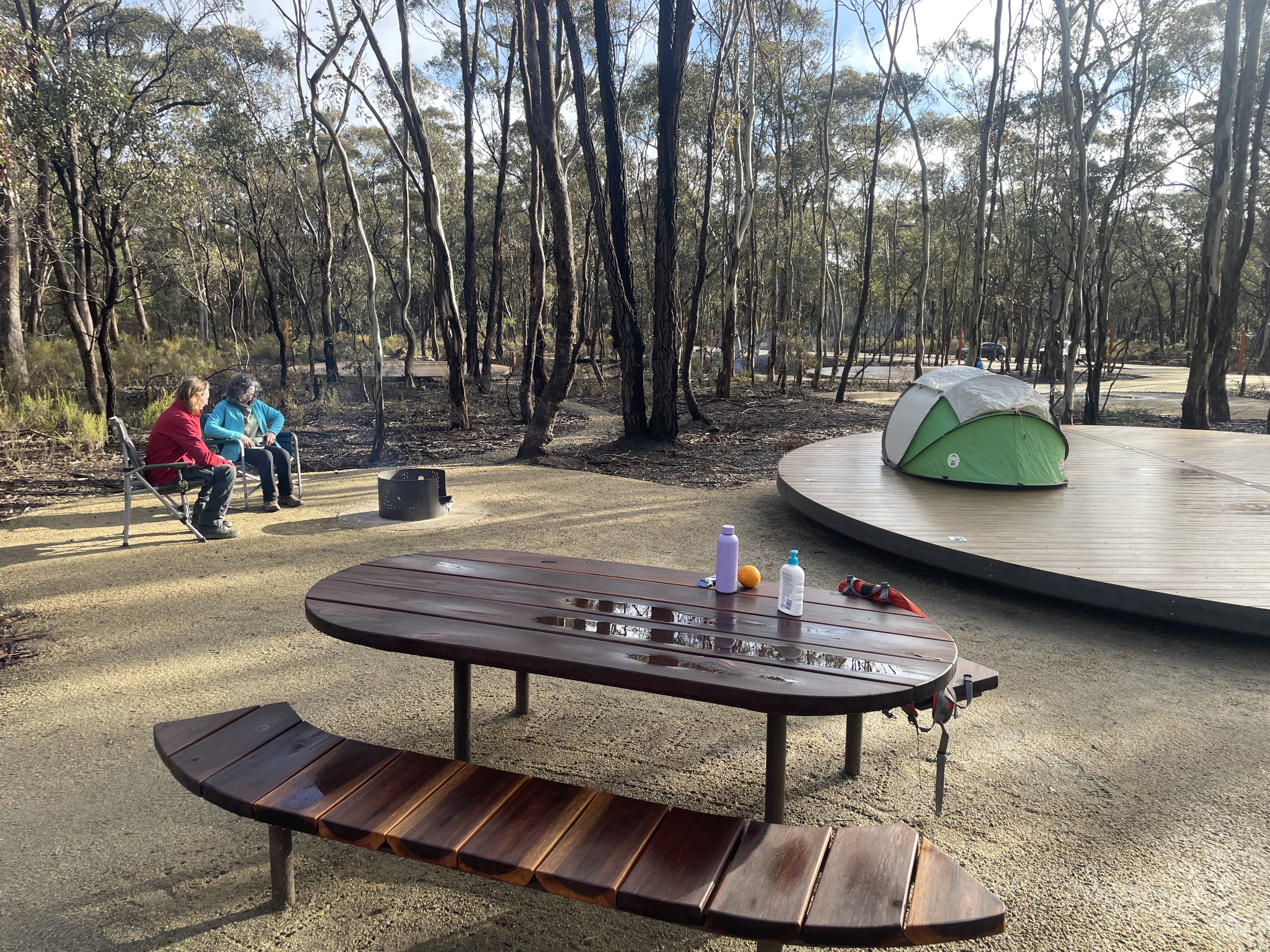 Campsite with table and chairs in foreground, tent and platform and two people sitting by fire in background