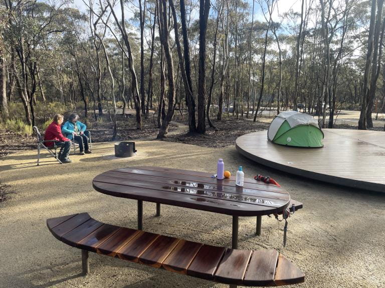 Campsite with table and chairs in foreground, tent and platform and two people sitting by fire in background