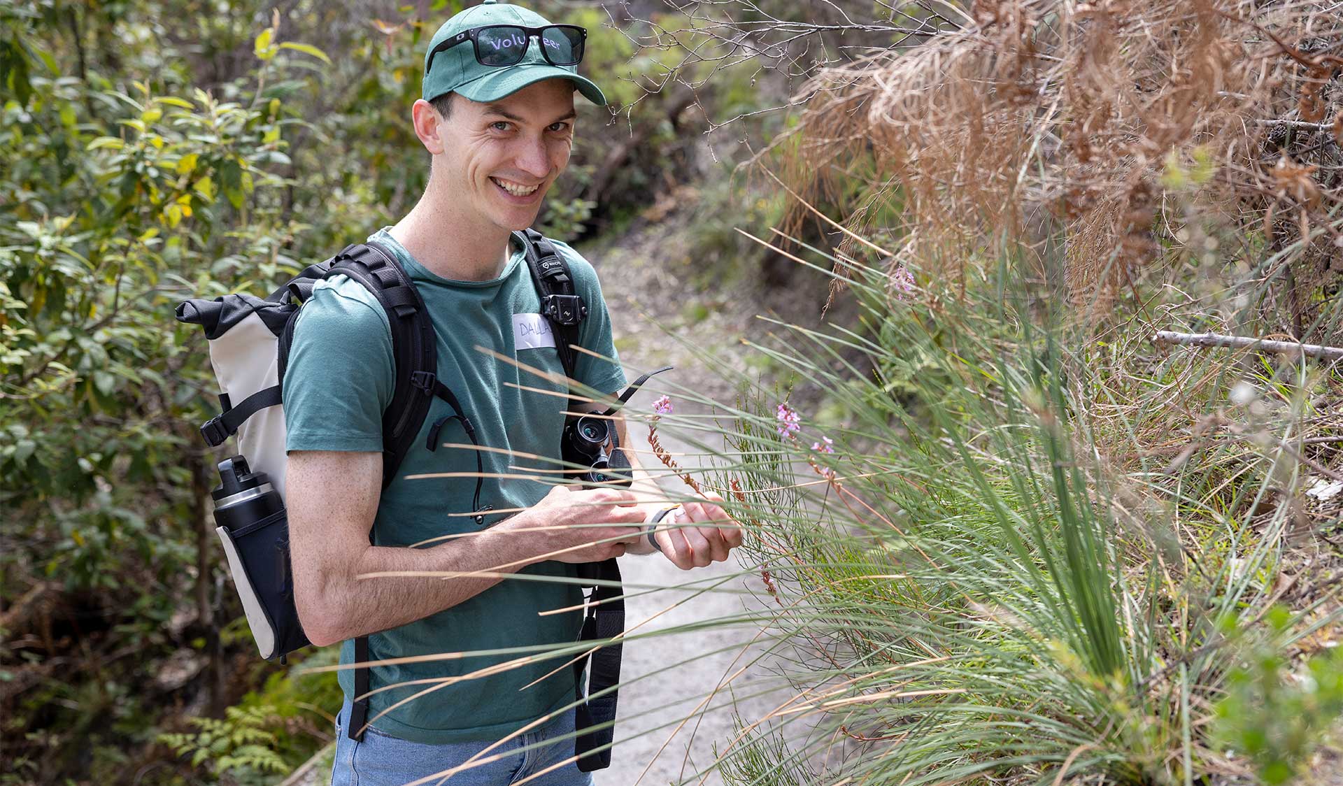 A volunteer dressed in a t-shirt, shorts, backpack, and baseball cap holding part of a plant while smiling at the camera. 