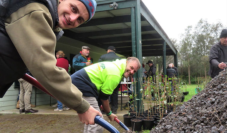 Two men smiling at the camera while shoveling a pile of gravel, with groups of people and tree saplings in the background.