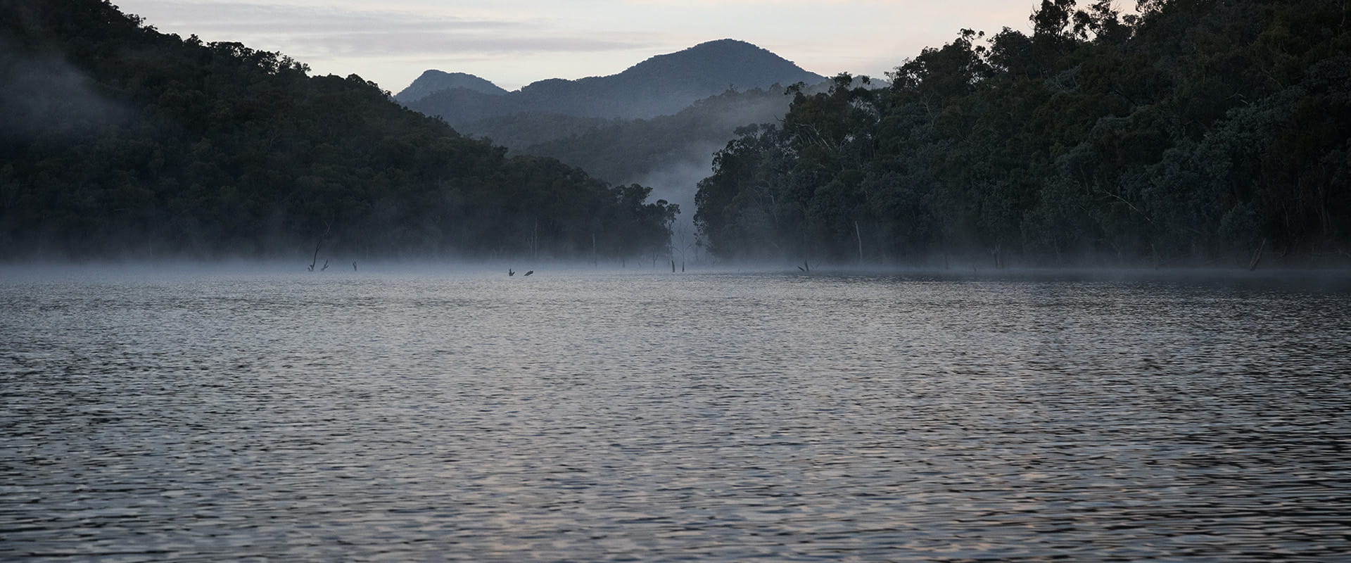 Mist rises on a lake in the foreground. Forested mountains rise in the background and far off in the distance. 