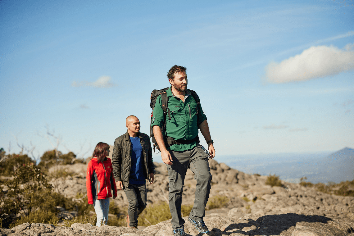 Three people hiking on rocky surfaces in the Grampians National Park with dense bushland, and a clear blue sky in the background.