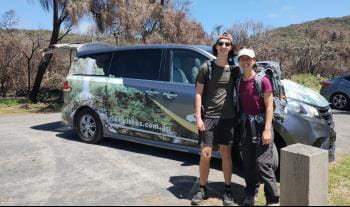 A smiling couple stands in front of a Ride With Us transport van.