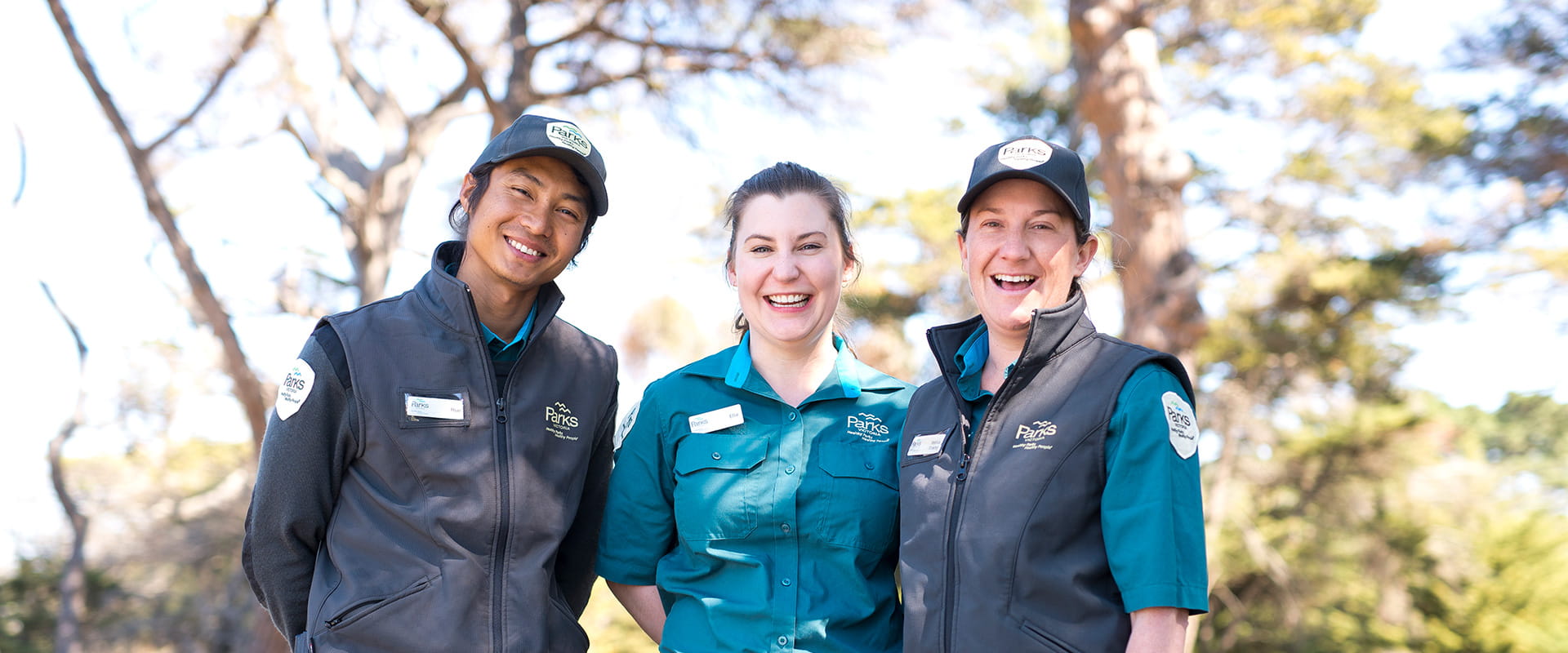 Three rangers in Parks Victoria uniform smiling at the camera with trees in the background.