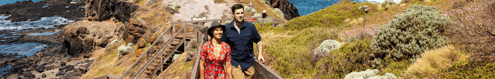 Two people walking along pathway with rocky landscapes in the background meeting the ocean.