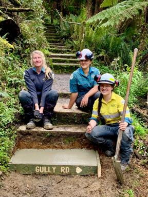 Shows a new concrete step in place on a freshly cleaned walking track surrounded by forest. Three women sit on the steps, one is a park ranger with working gloves and two others are in high viz with overalls and hard hats, all are smiling at the camera with a sense of a job well done.
