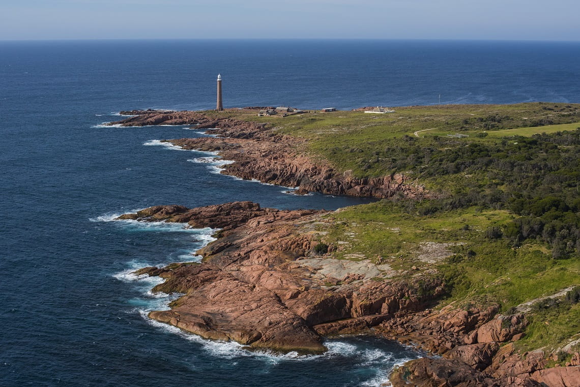Gabo Island from above. A rocky coastline meets the blue ocean, with green foliage covering the island towards the centre. A lighthouse is perched on a rocky outcrop in the distance.