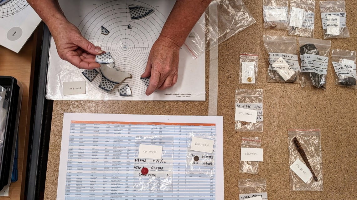 A table with pieces of pottery in small bags: a pair of hands are holding some of them, placing them on a piece of paper marked with the outline of a plate.