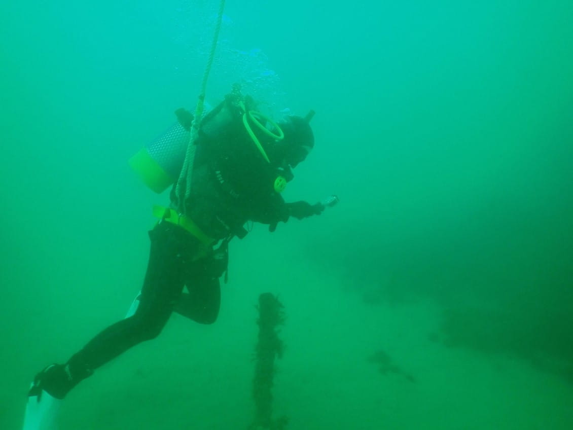 A person in scuba diving gear, underwater, holds a magnifying glass to look at a rusting metal bar.
