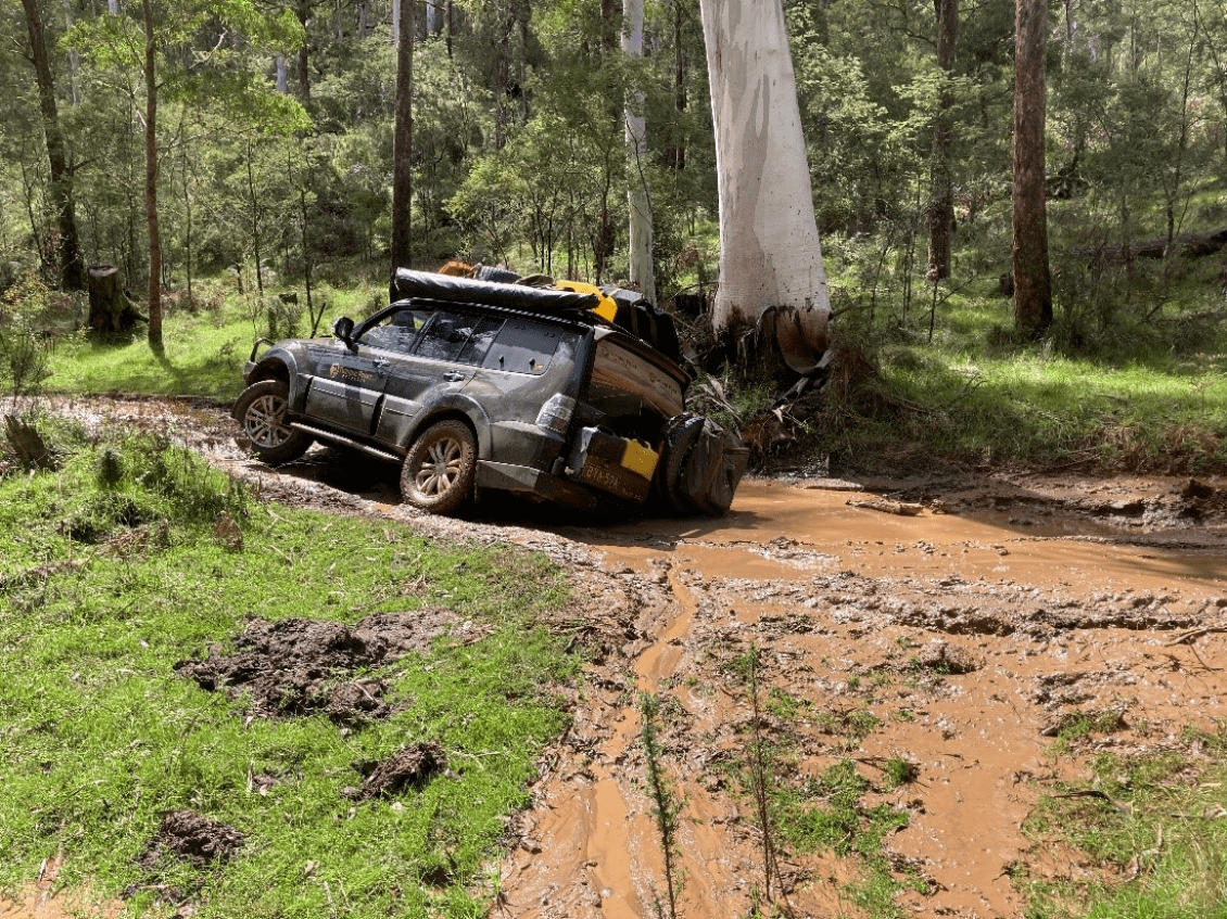 A four wheel drive tipped on its side, stuck in flood waters on an unpaved road.