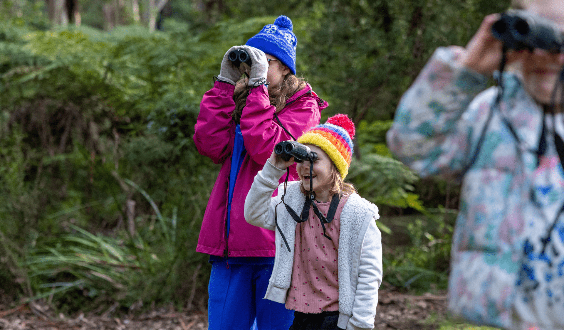  Three children wearing bright jackets and beanies look through binoculars in a grassy clearing, birdwatching in a park.