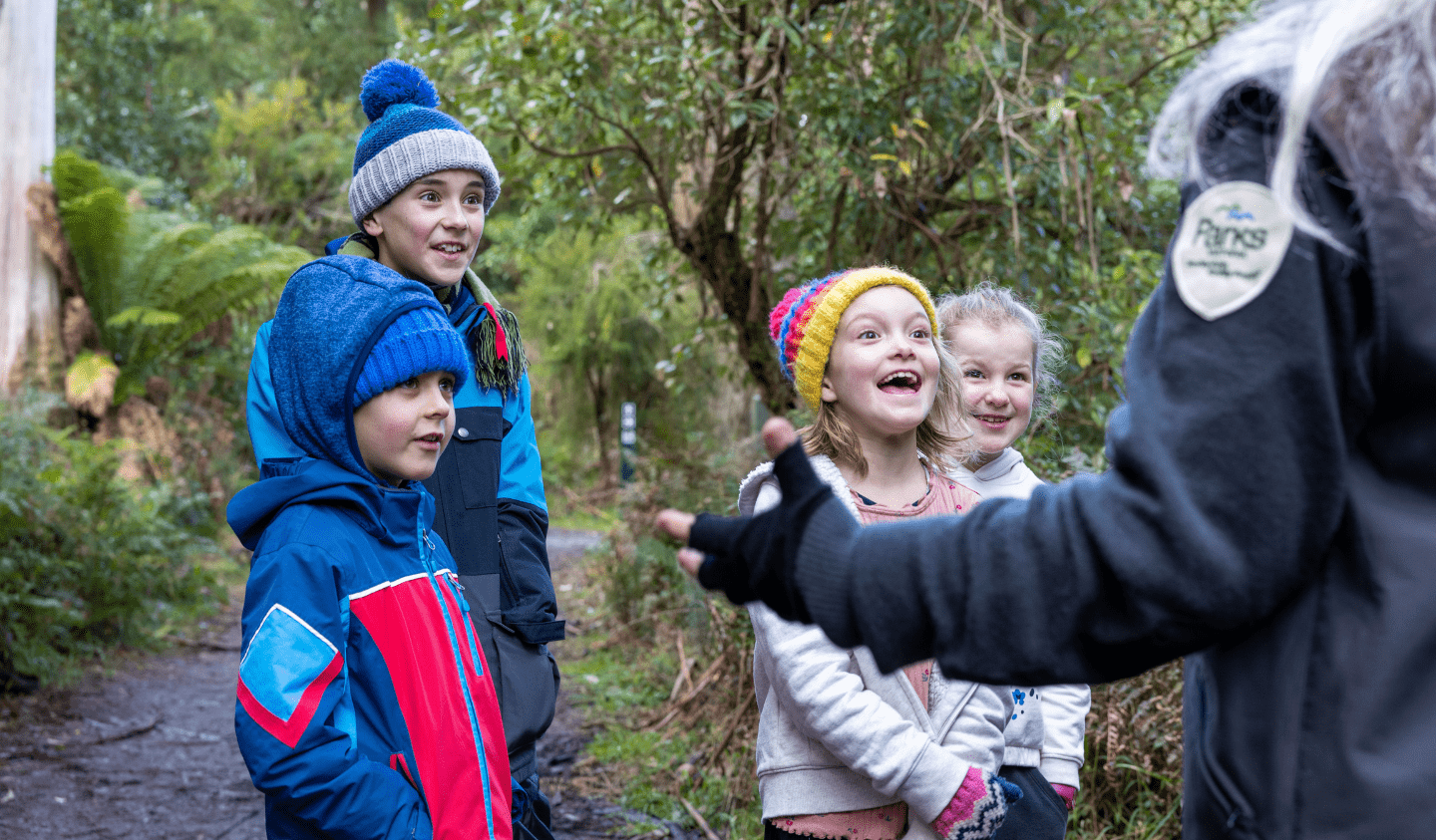 Four children in warm jackets and beanies smile and listen attentively to a ranger on a bush track in a green forest setting.