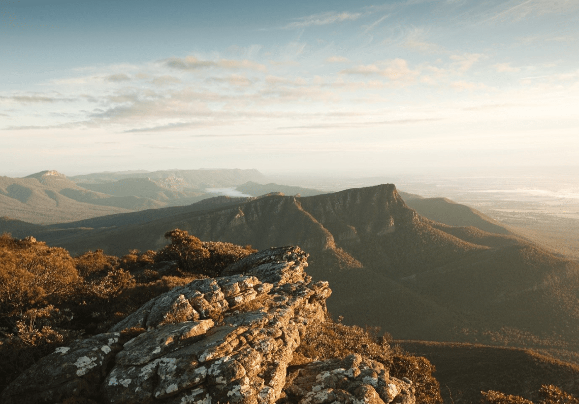 The mountainous landscape of Gariwerd is seen from an expansive distance, with rocks in the forefront and forested areas immediately behind it.