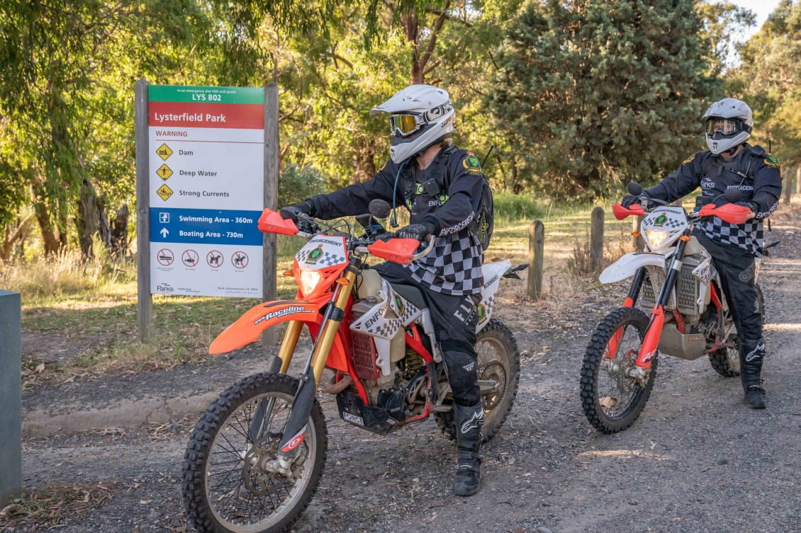 Two people on trailbikes ride past a sign saying "Lysterfield Park"