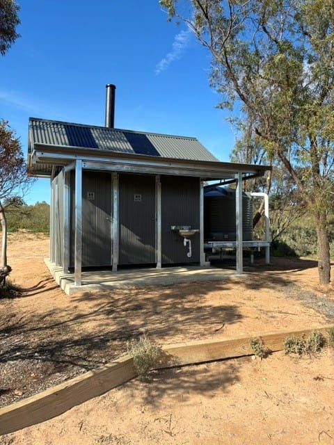 Grey metal two cubicle toilet block with a water tank surrounded by trees