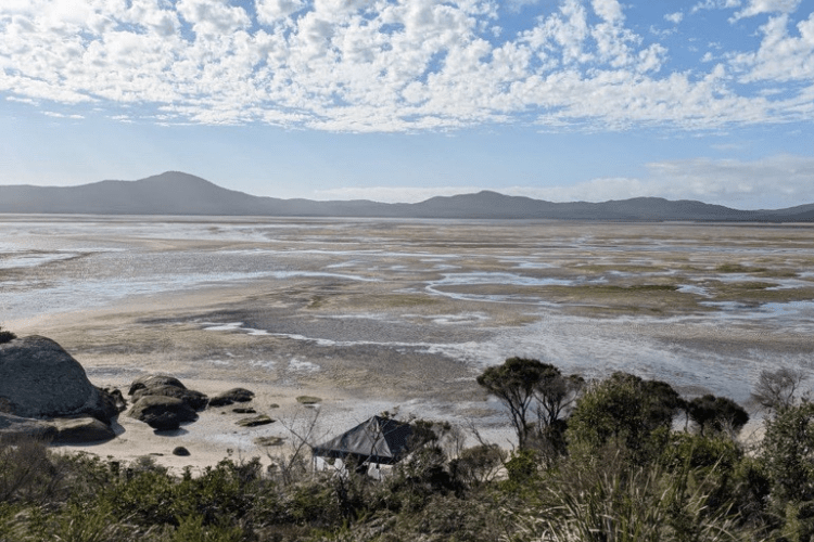Wide coastal landscape showing an expansive tidal flat at low tide, patterned with shallow water channels and patches of exposed sand and mud. In the foreground, a slope with green shrubs and grasses leads down to scattered rocks and a small structure with a pitched roof. In the distance, low, hazy mountains stretch across the horizon beneath a blue sky filled with scattered, wispy clouds.