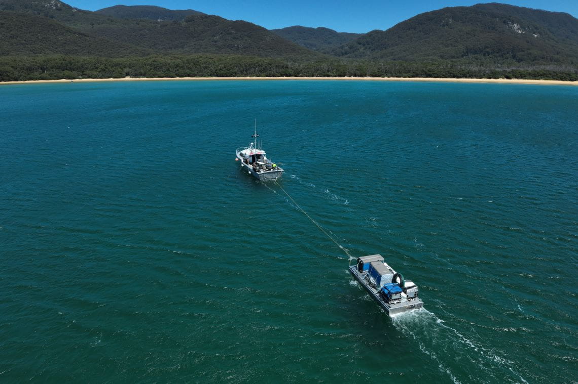 A grey boat tows a barge towards a long beach, with mountains behind. The barge is full of objects covered with blue tarpaulin,
