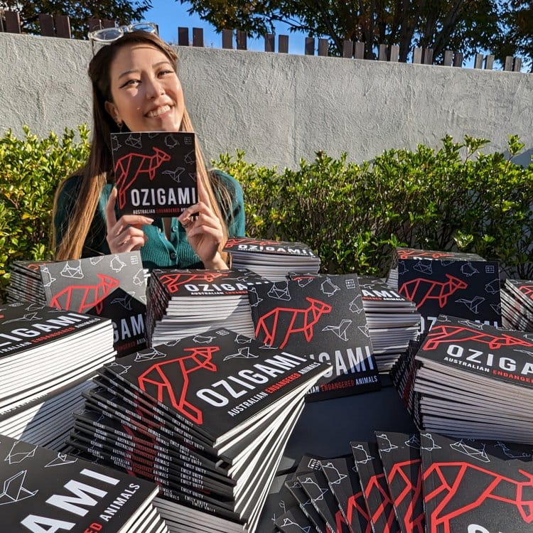 Emily sits in front of a pile of origami books, she holds one up for the camera