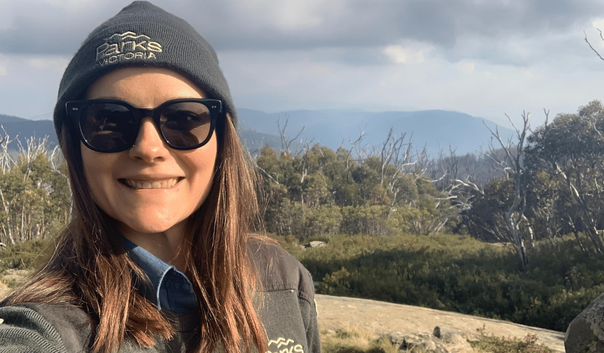 Mel standing in her Parks Victoria uniform, smiling proudly with a view of the Yarra Ranges in the background