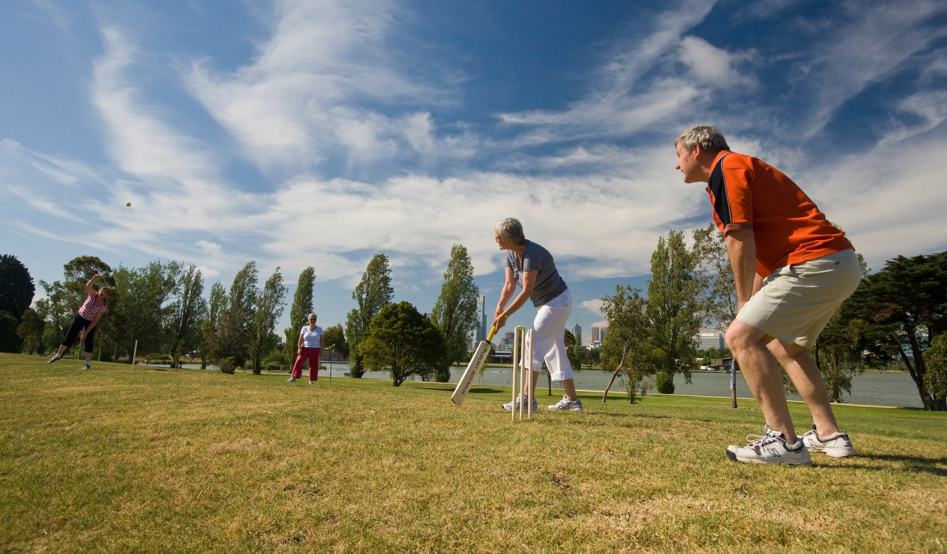 A group of friends play a casual game of cricket on the lawn, with Albert Park Lake in the background