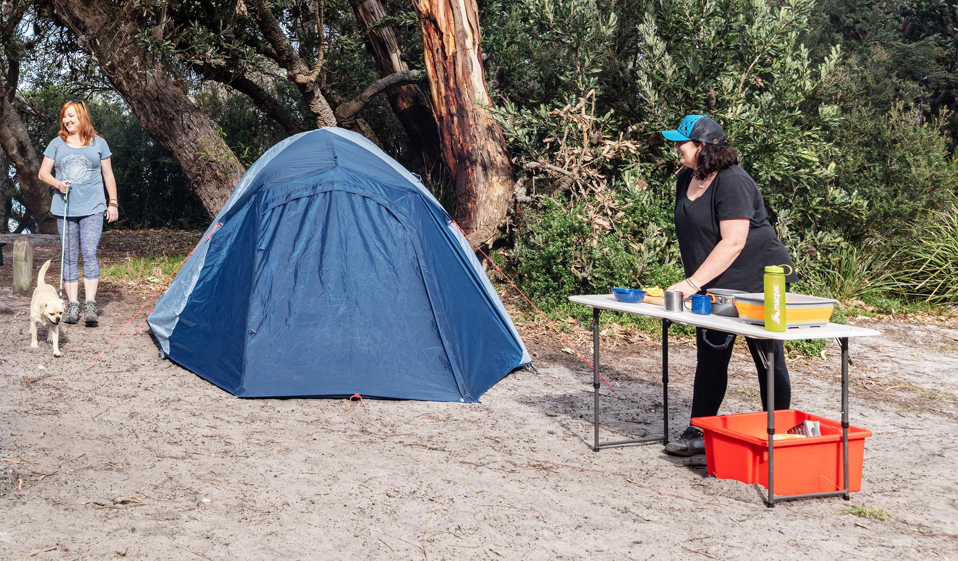 A women walks her dog into the campsite while another women prepares food as her son looks on.