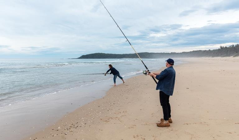 Two men cast fishing lines on a beach.