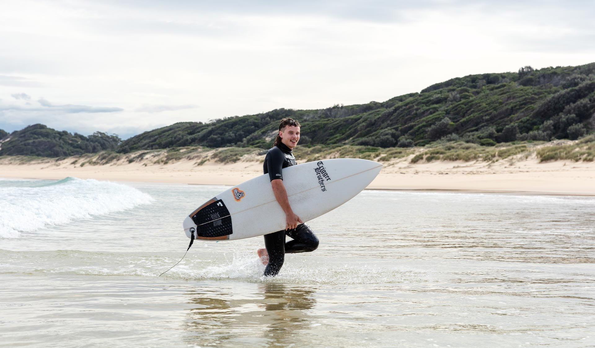 A man carrying a surfboard walks through the shallow water at West Cape Beach at Cape Conran Coastal Park