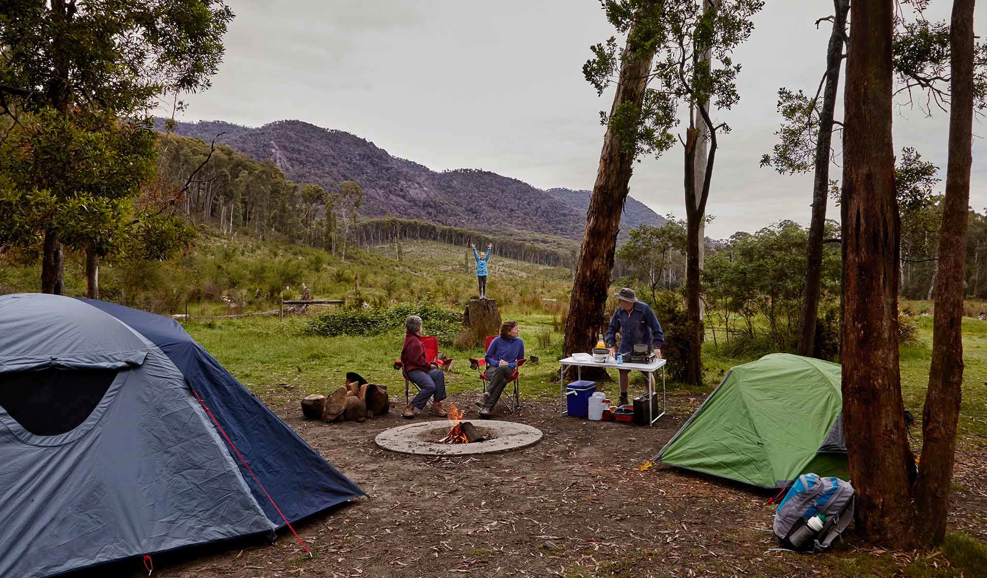 Two Grandparents and a mother sit by the camp fire while a young child plays in the background.