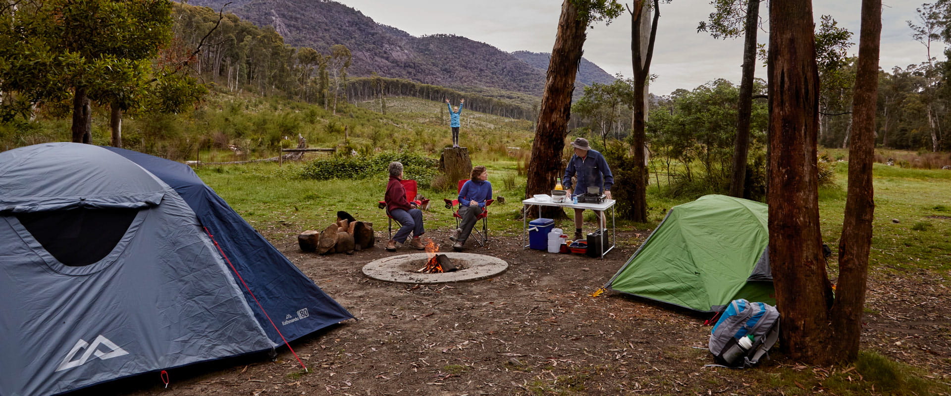 A woman and man talk to one another next to a fire pit at their campsite, while a woman sits and looks back at a young girl stretching her arms to the sky as she stands on a large stump with the mountains in the distance behind her.