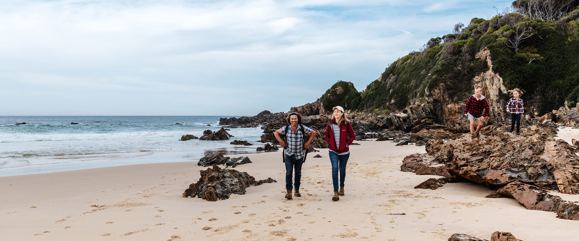 A father and moth walk along a sandy beach while their two children climb on the nearby rocks