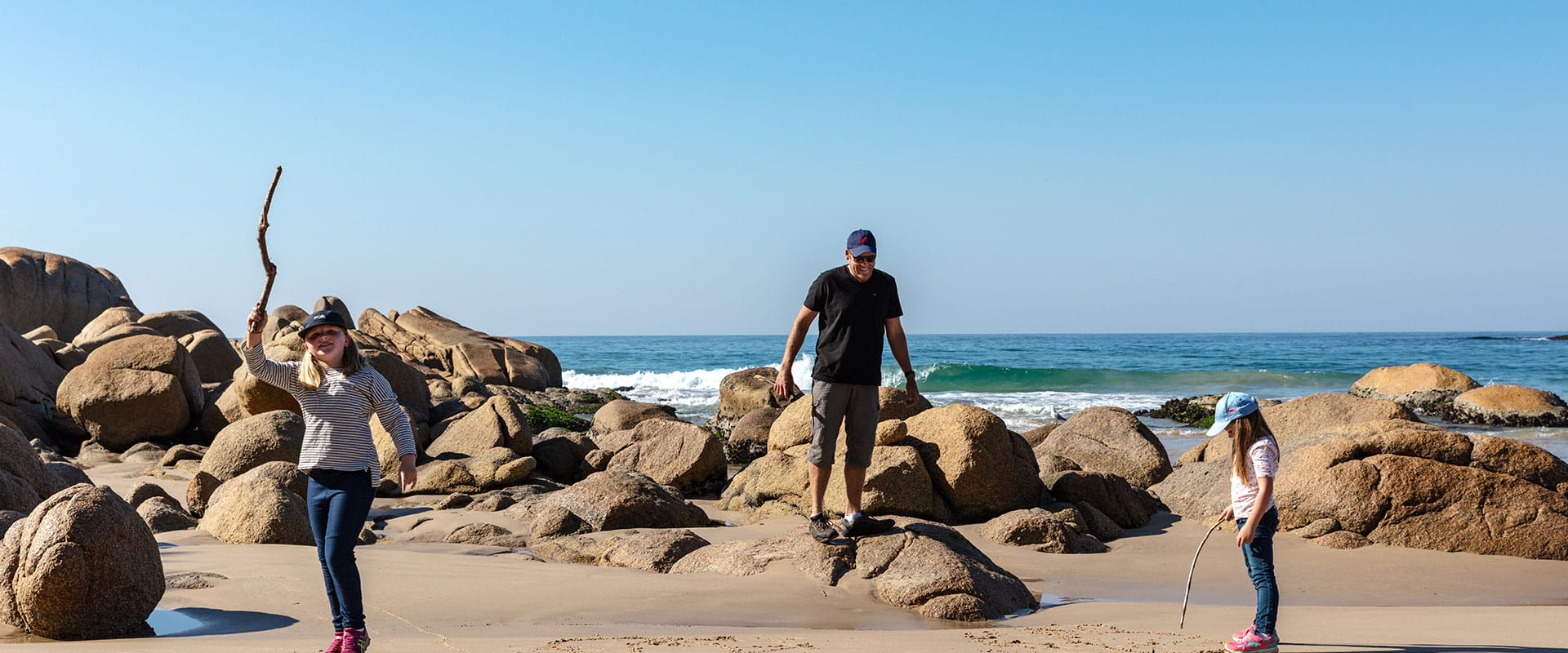 A father and his two daughters explore granite boulders on a sandy beach