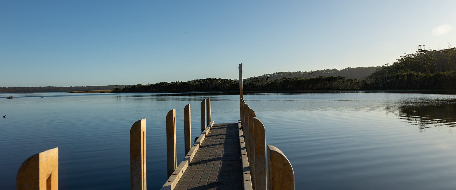 A pier leads out to a perfectly still blue lake in a brilliantly blue sky in the background.