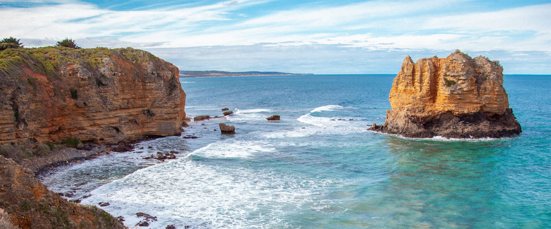 A rocky outcrop rises from the ocean, offering a clifftop view of long beaches and dunes in the distance.