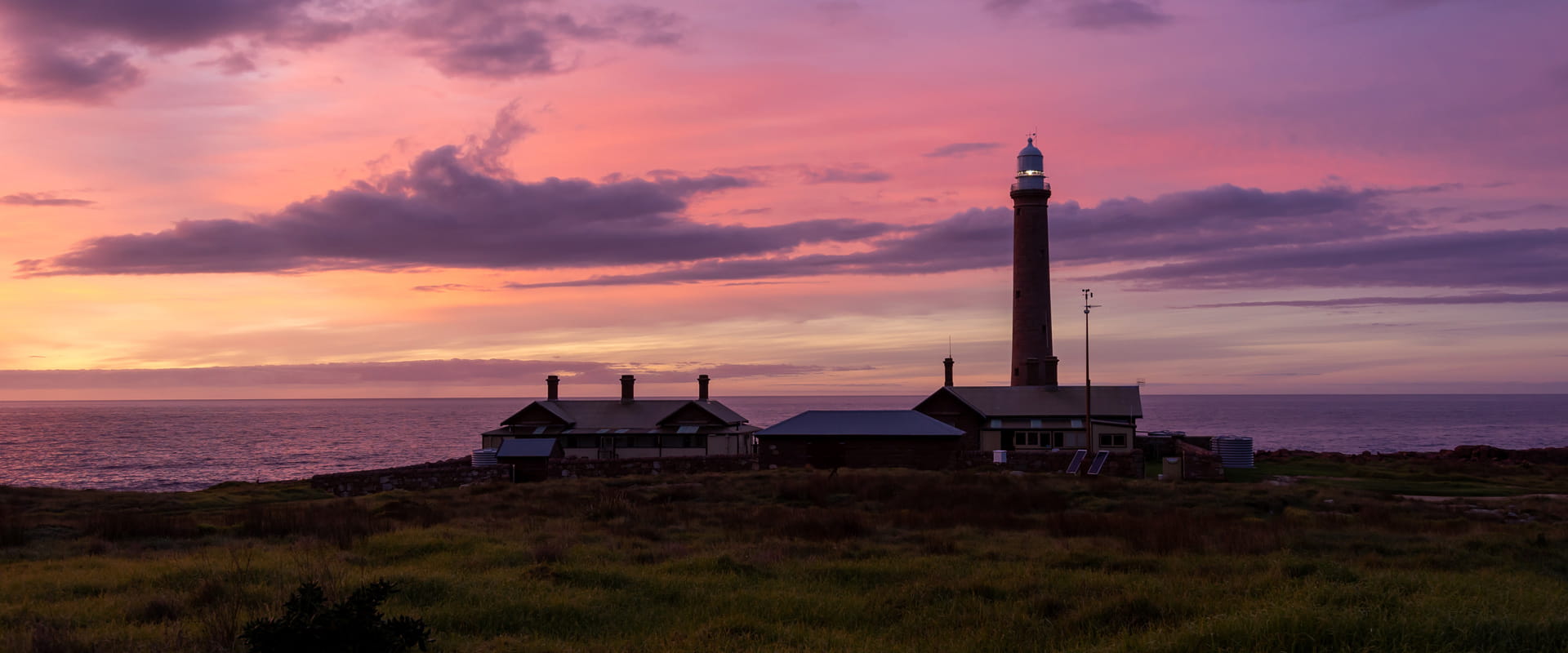 A dimly lit lighthouse perched on a cliffside overlooks the open waters as the sun sets against a purple sky.