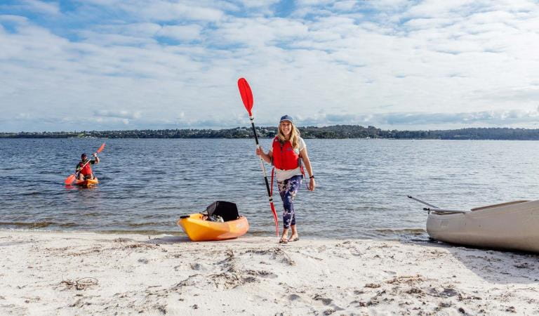 A woman holding a paddle walks up the beach with her kayak on the beach behind her
