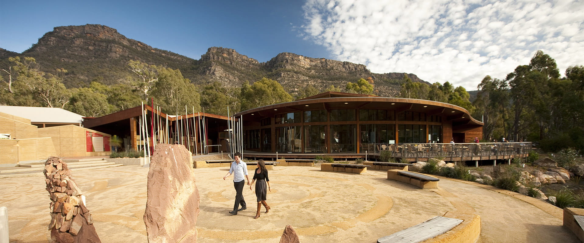 A man and a woman walk past the Brambuk building with mountain peaks in the background.