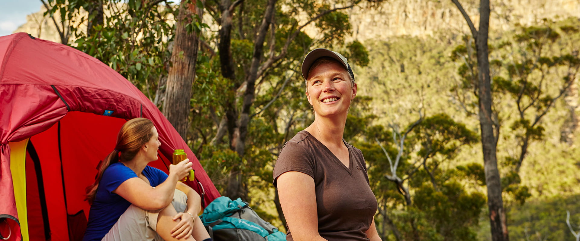 Two hikers sit outside of their tent on a wooden tent pad enjoying a drink
