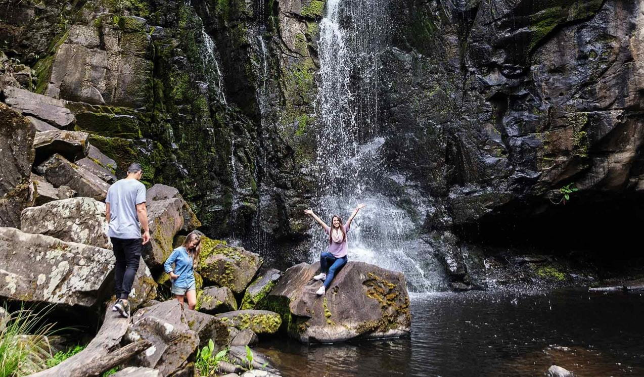 A women sitting on a rock in front of a waterfall calls out to her friends to join her.