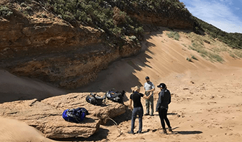 Three hikers with their packs on a rock at the beach along Great Ocean Walk