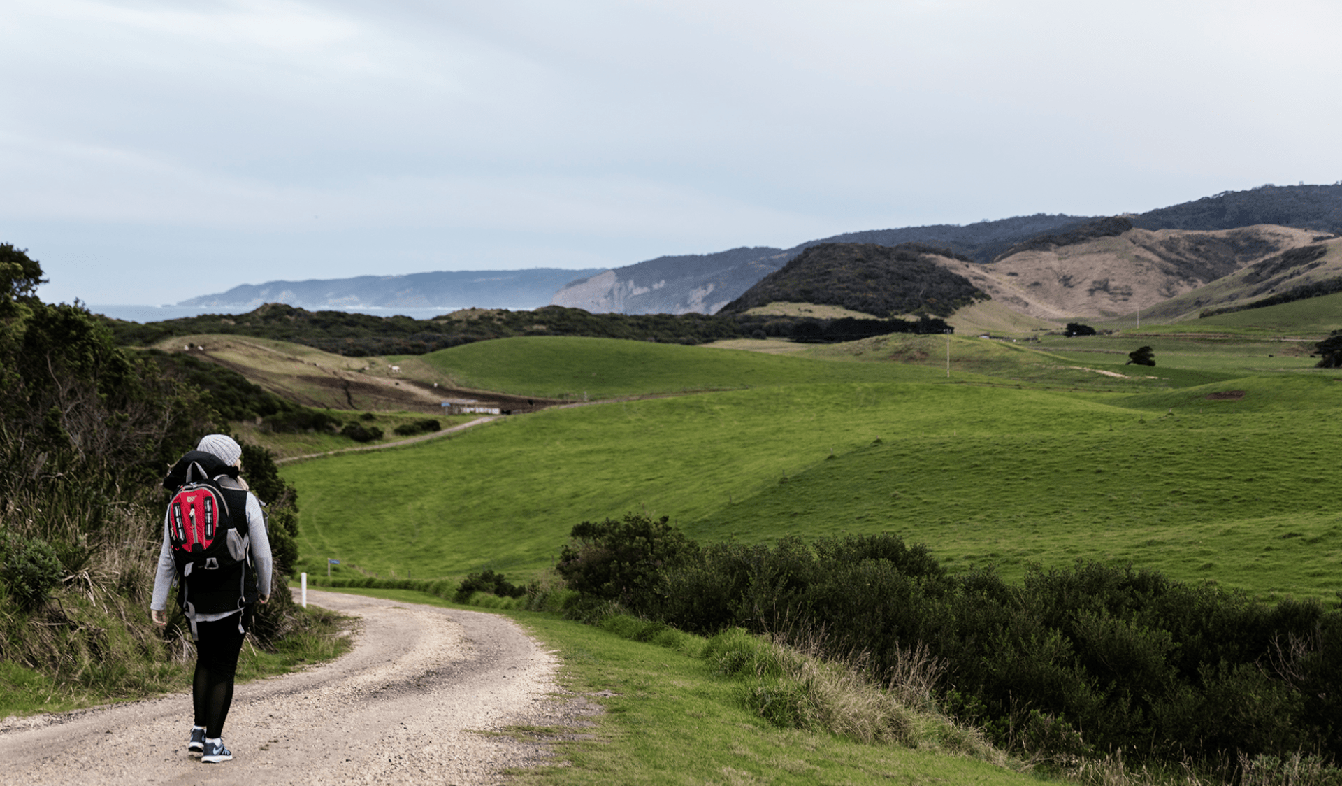 A woman with a small backpack walks along a road surrounded by hills