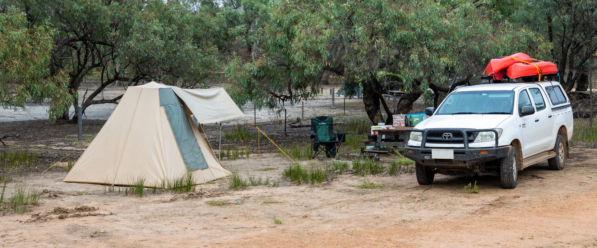 A camping tent and folding chair set up next to a trailer with food supplies parked by a wooden picnic table under the shade of a nearby tree.
