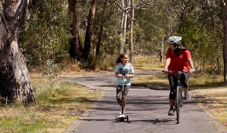 A woman riding a bike alongside a young girl on a scooter